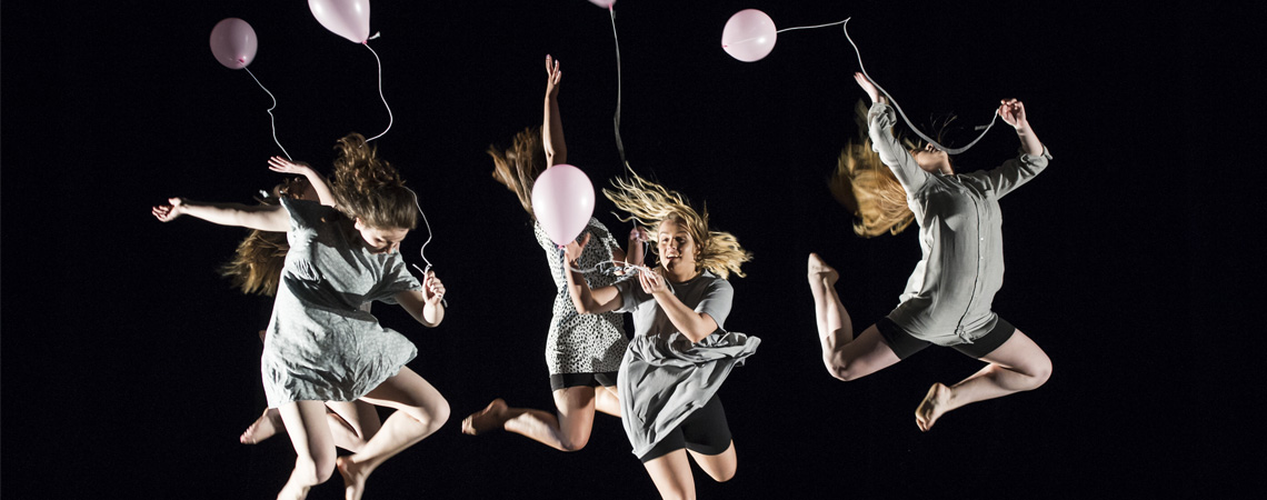 A group of dance students on stage holding pink balloons and jumping into the air as part of a performance