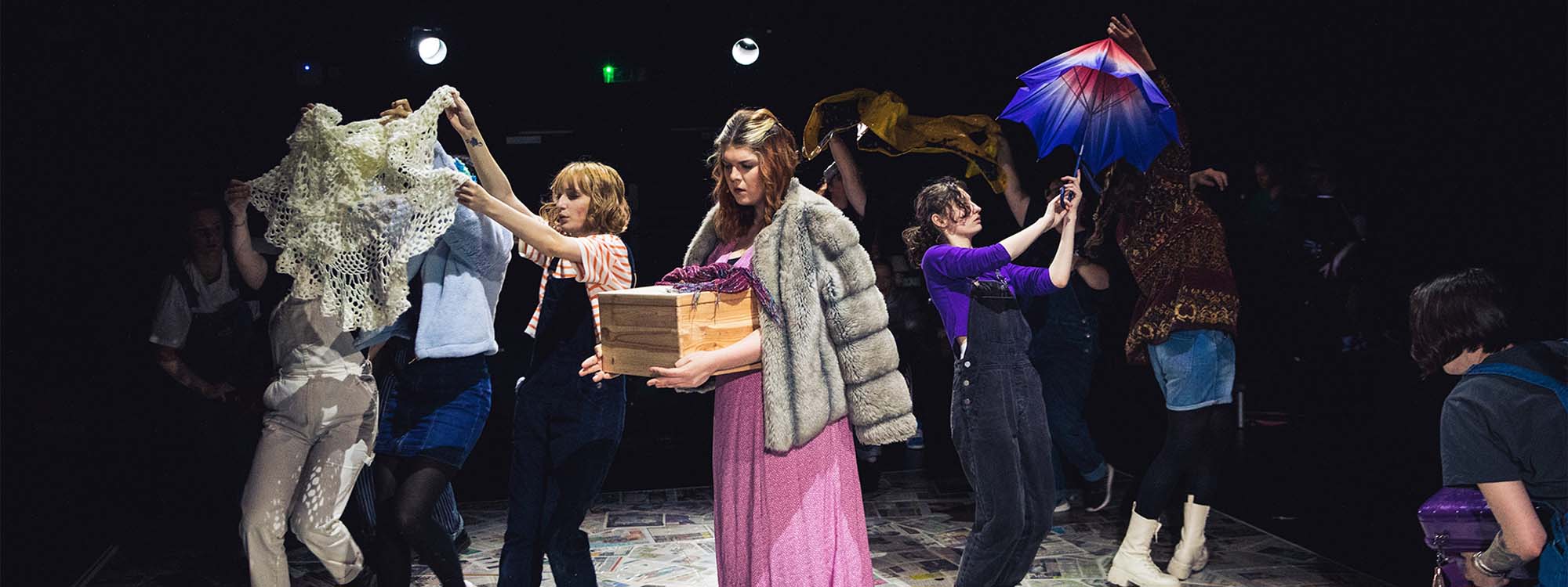 Students performing on stage holding objects such as a wooden box, cardigan, and umbrella