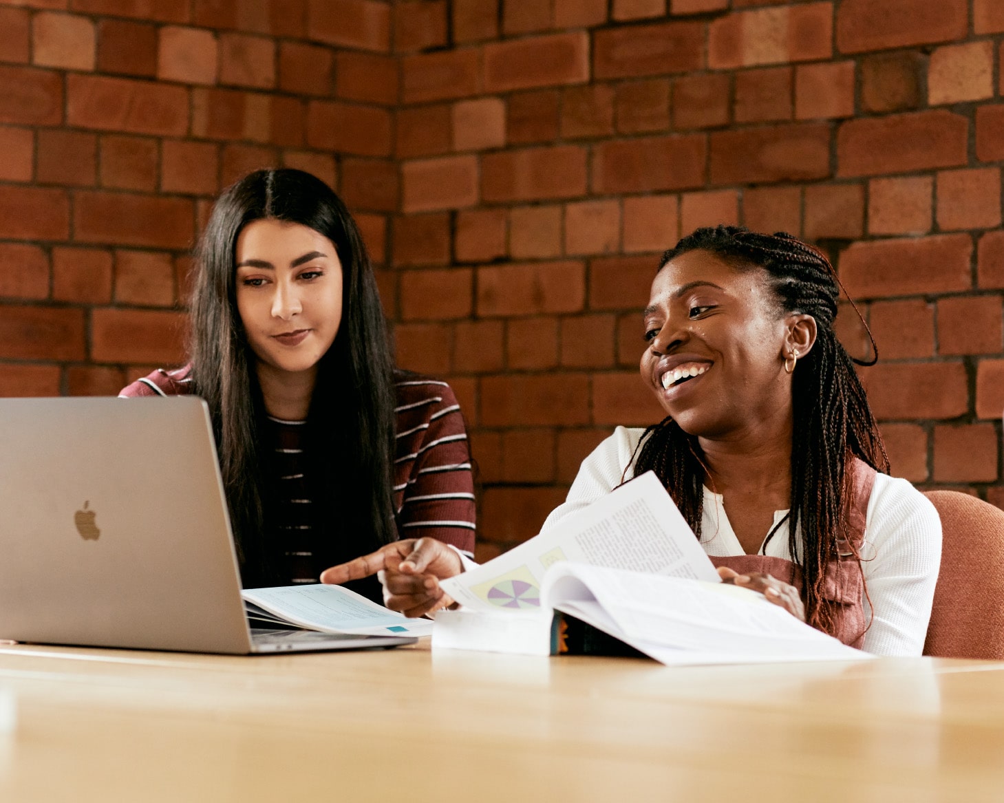 Two female students sat in the library, looking at a laptop and a textbook