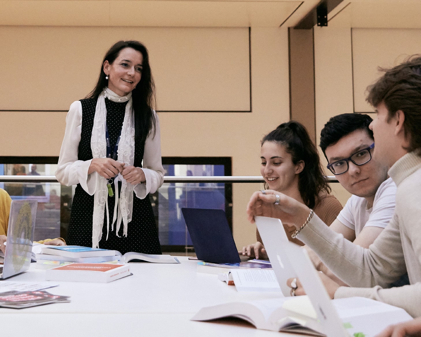 A staff member teaching a group of students who are sat at a table with an array of textbooks and laptops on